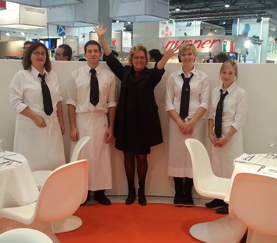 Frauke Schellhas steht zwischen vier Mitarbeitern, Alle lächeln in die Kamera, Mitarbeiter tragen weiße Hemden, Blusen und Schürzen, schwarze Krawatte mit Logo der Firma Schellhas Event & Catering